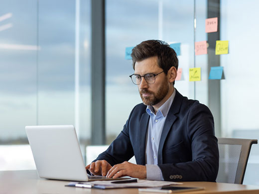 Focused businessman concentrates on his laptop at a well-organized office desk with sticky notes in the background.